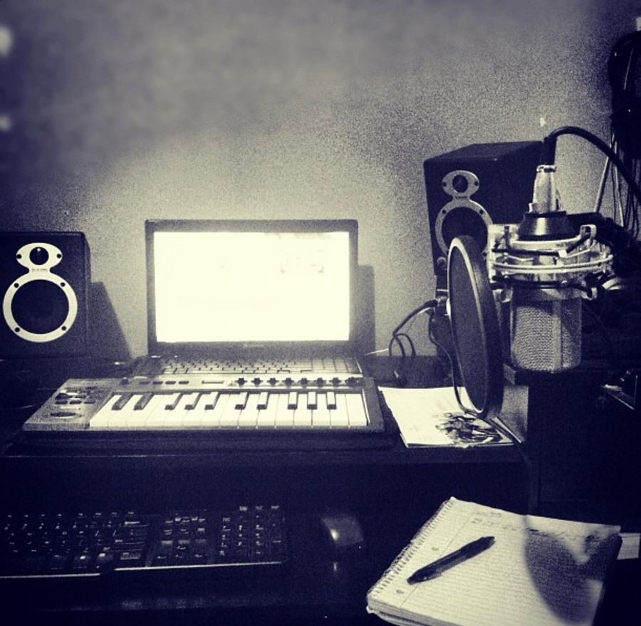 Early home studio desk in black and white: laptop, MIDI keyboard, two monitors, a condenser mic with pop filter and a notebook